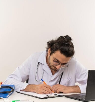 Doctor focusing on paperwork and documentation with a laptop on the desk, highlighting the challenges of medical documentation.