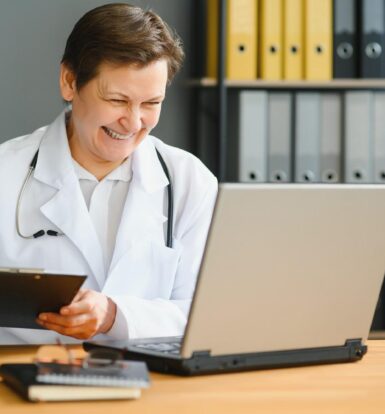 Happy doctor using a laptop while taking notes, representing the efficiency of Virtual Medical Receptionist Services.