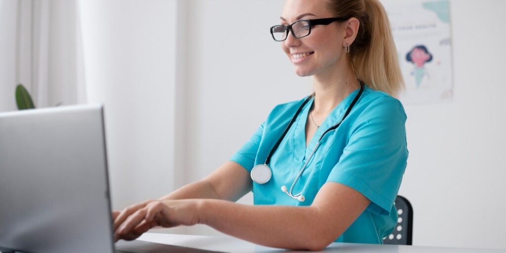 Smiling female Virtual Medical Assistant in blue scrubs working on a laptop, managing healthcare administrative tasks.