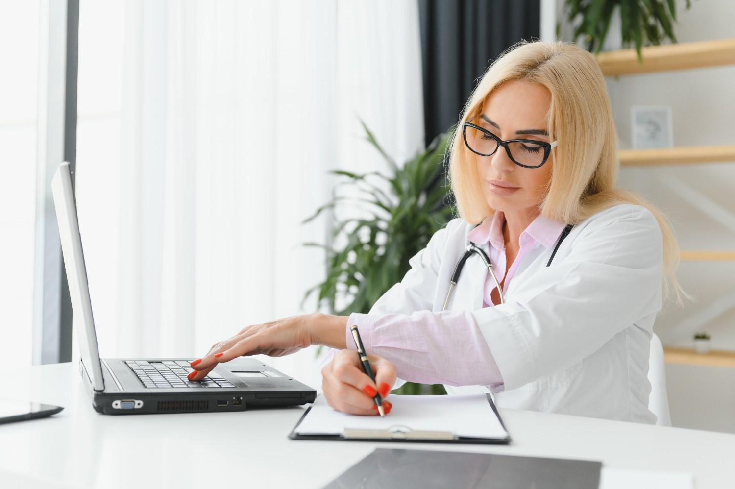 Doctor verifying insurance details on a laptop while taking notes.